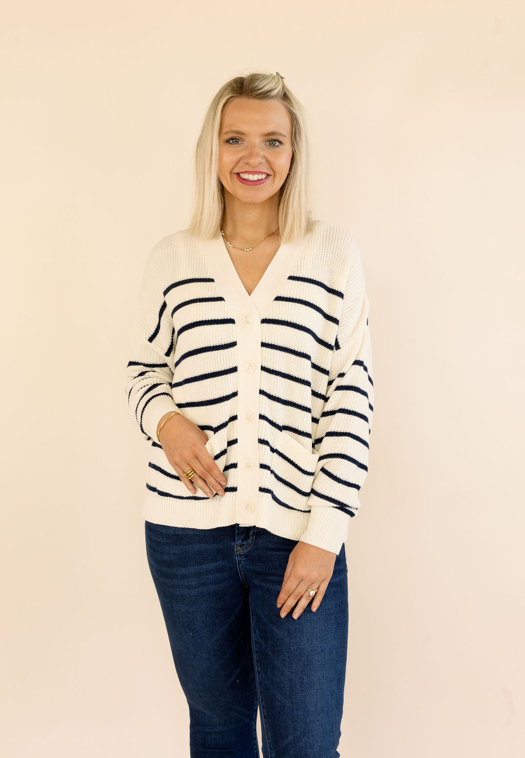 A smiling blonde woman wearing a cream-colored knit cardigan with navy blue horizontal stripes and dark blue jeans, standing against a solid light beige background.