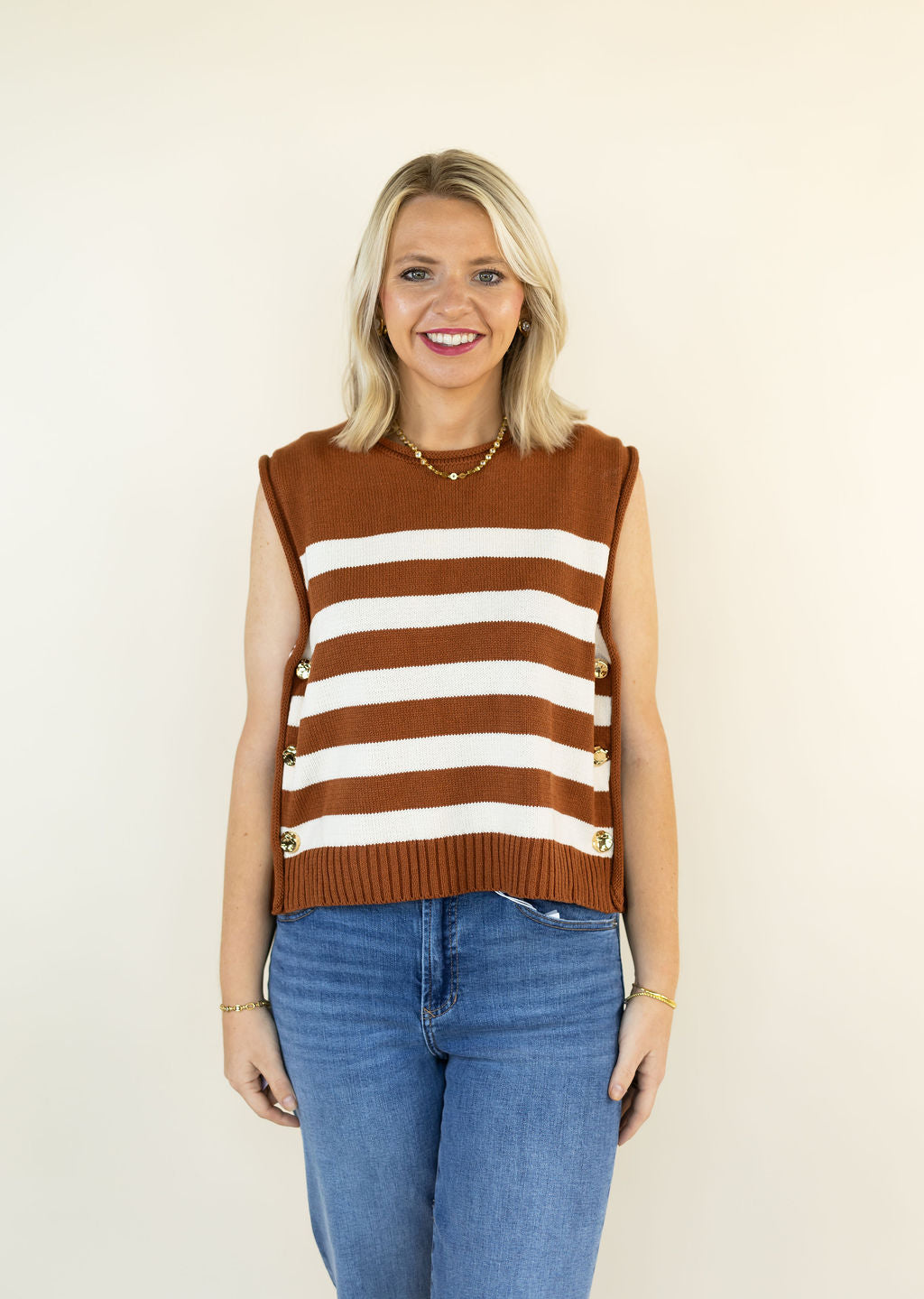 A smiling blonde woman wearing a sleeveless caramel and white striped knit sweater vest with gold-tone decorative buttons on the sides, paired with blue jeans, standing against a plain cream background.