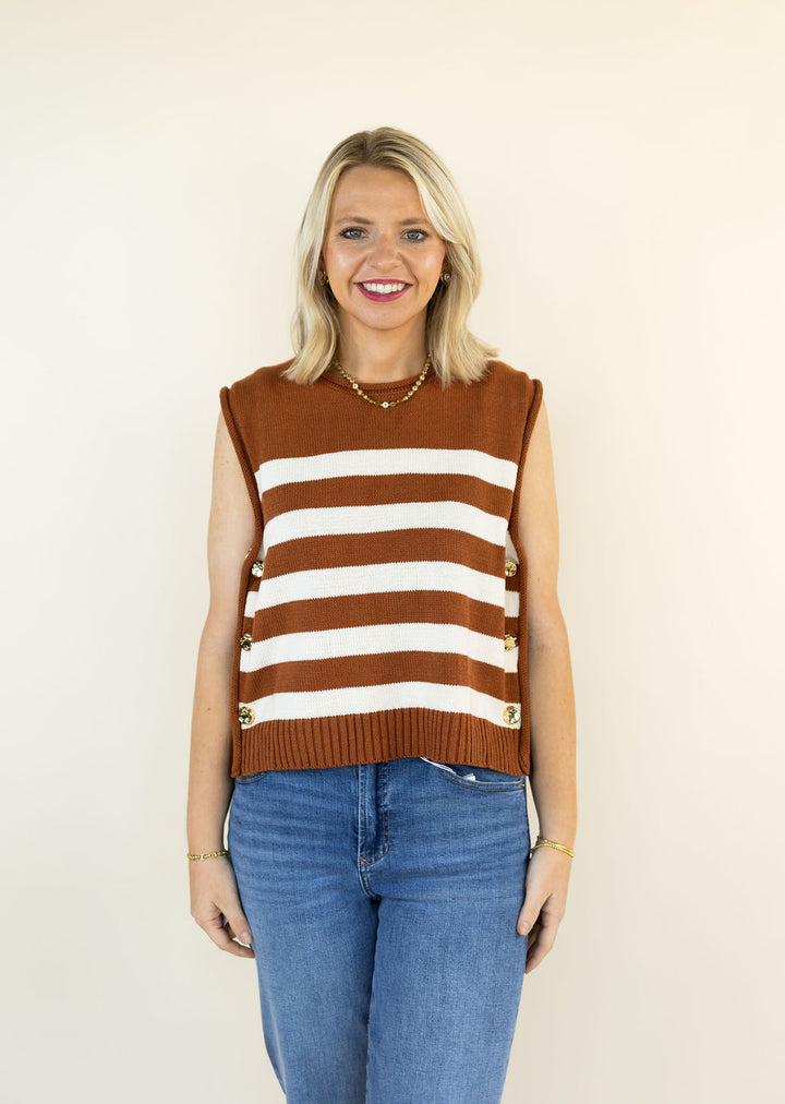 A smiling blonde woman wearing a sleeveless caramel and white striped knit sweater vest with gold-tone decorative buttons on the sides, paired with blue jeans, standing against a plain cream background.