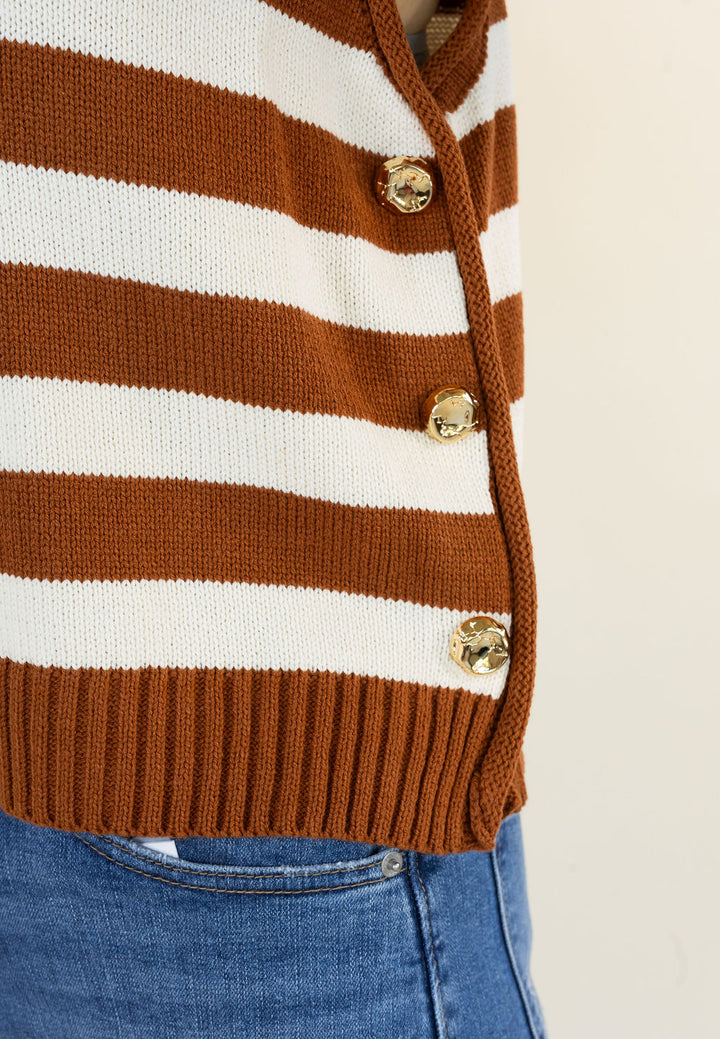 Close-up of a caramel and white striped knit cardigan with gold buttons, worn with blue jeans against a light beige background.