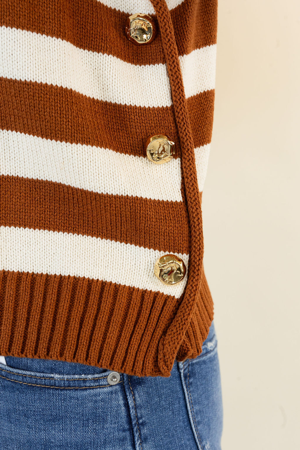 Close-up of a caramel and white striped knit cardigan with gold-tone buttons, worn over blue jeans against a plain light background.