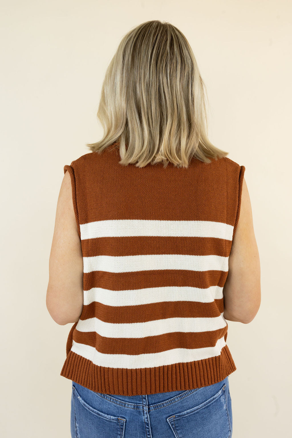 A woman viewed from behind wearing a sleeveless knit sweater vest with horizontal caramel and white stripes, paired with blue jeans, against a plain beige background.