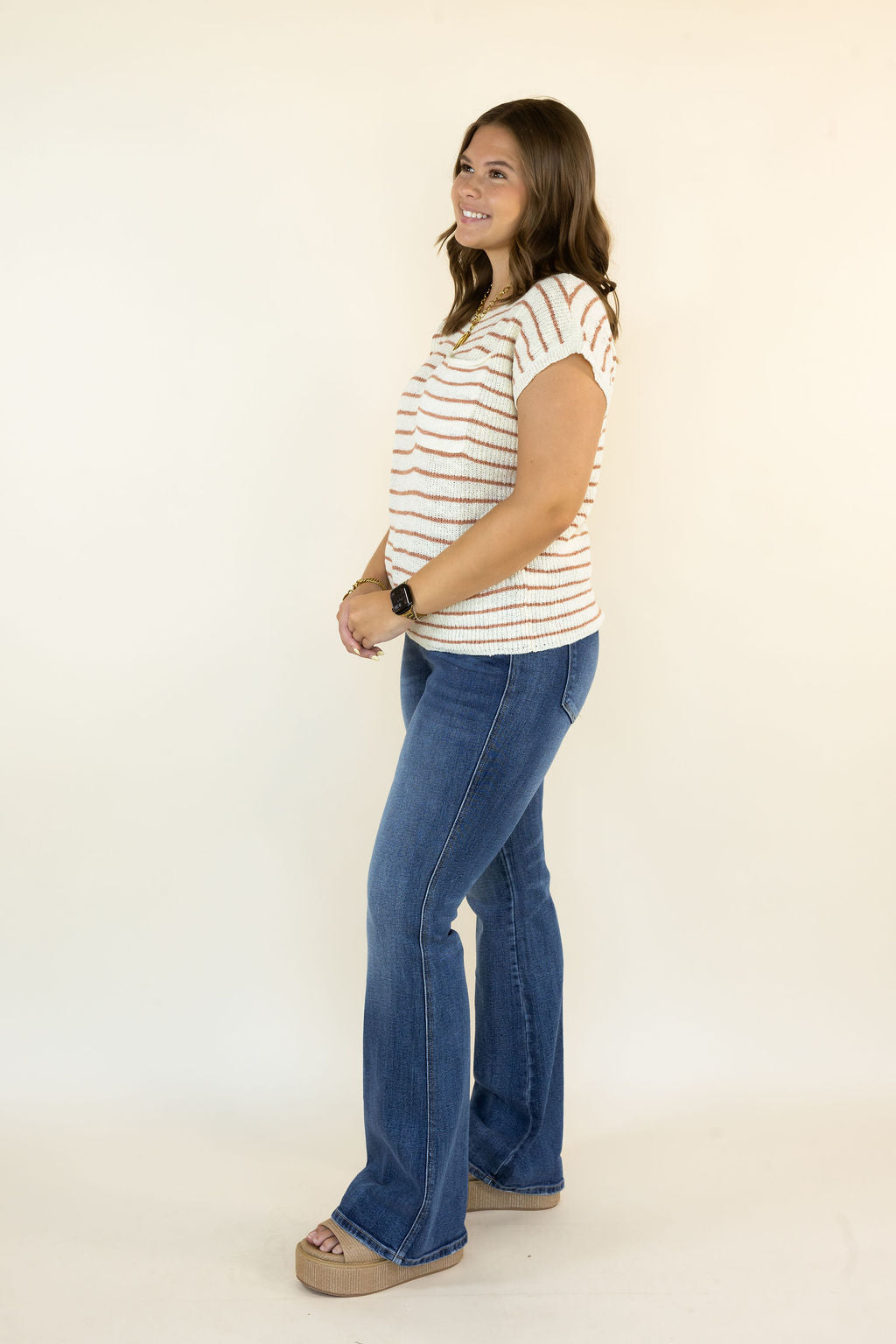 A woman modeling a short-sleeve crewneck t-shirt with thin orange and white horizontal stripes, paired with blue flare jeans and tan platform sandals, standing against a plain off-white studio background.