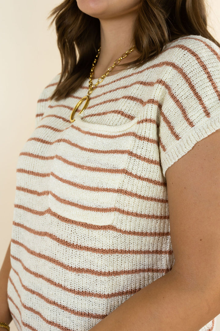 A close-up of a woman wearing a cream knit short-sleeve top with thin rust-brown horizontal stripes and a gold pendant necklace.