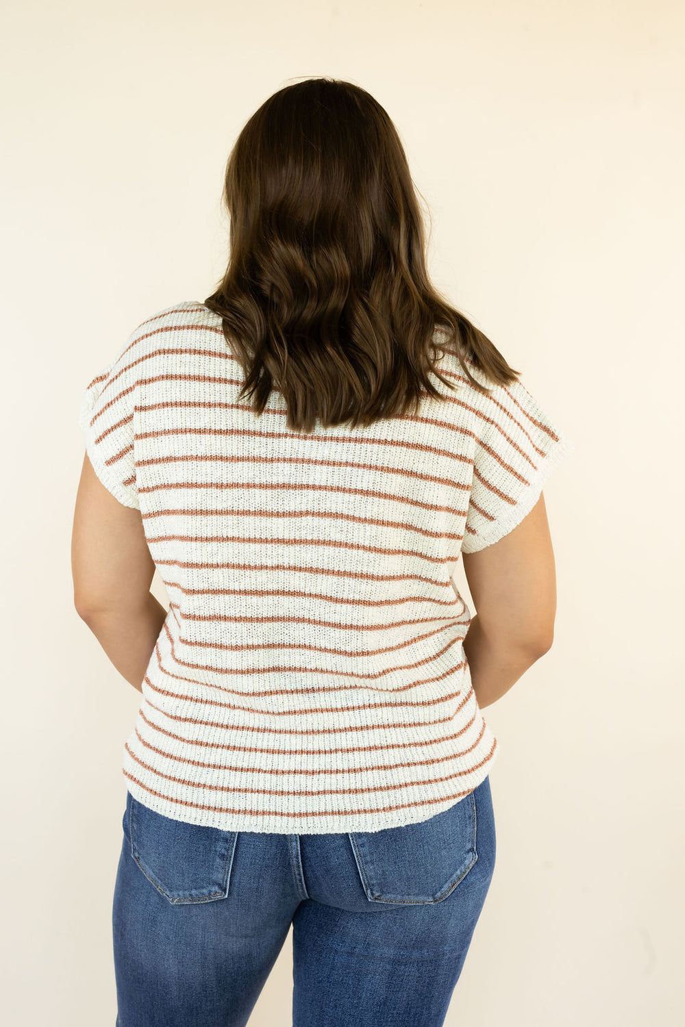A woman viewed from behind wearing a cream-colored short-sleeve top with thin brown horizontal stripes and blue jeans, standing against a plain off-white background.