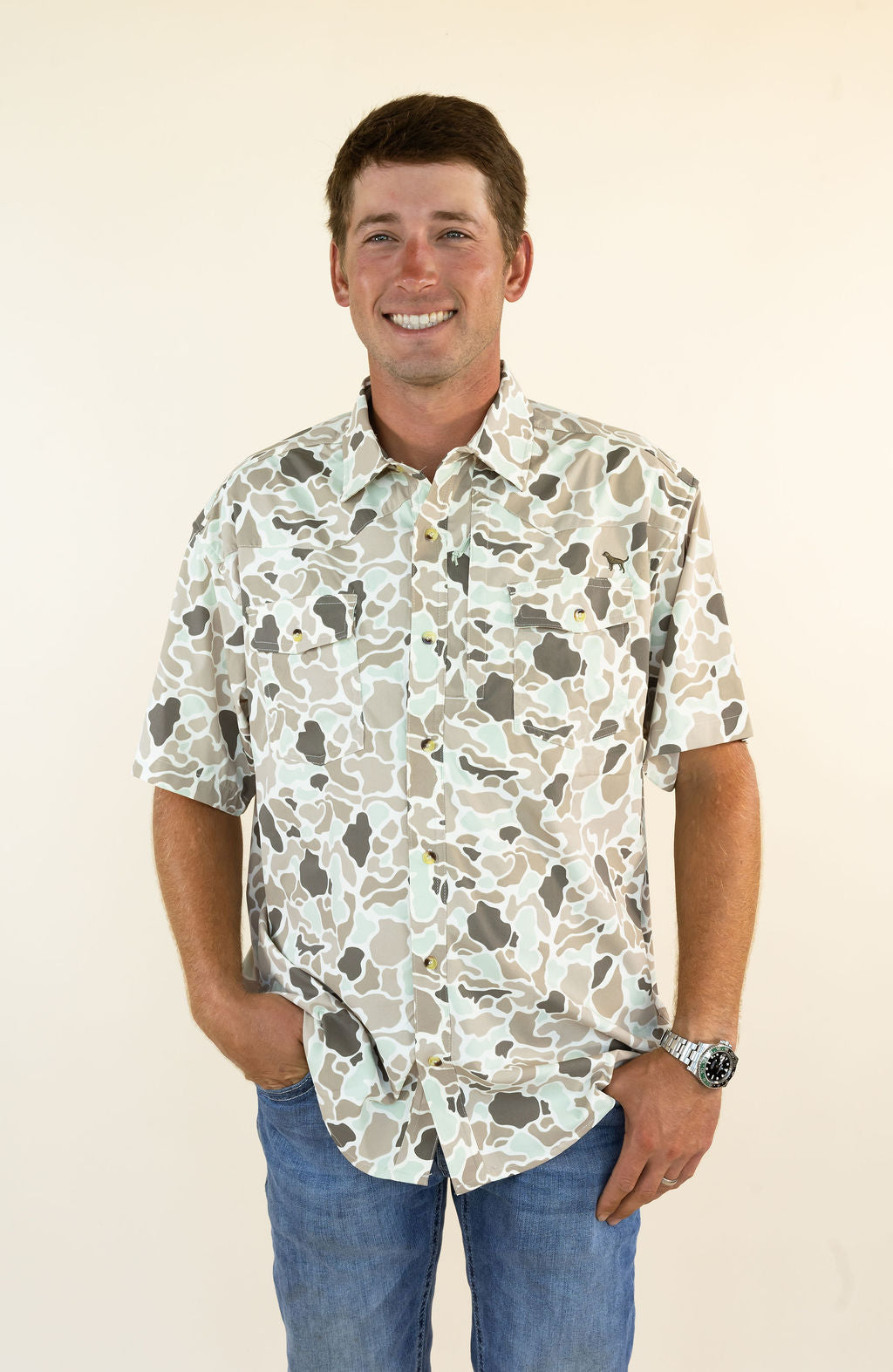 A smiling man wearing a short-sleeve button-up shirt with a beige, gray, and white camouflage pattern, paired with blue jeans, standing against a plain cream background.