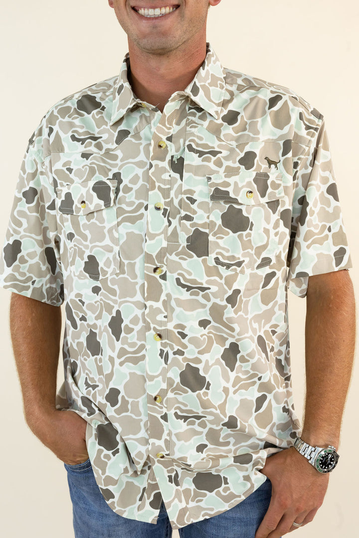 A man wearing a short-sleeve button-up shirt with a beige, gray, and off-white camouflage pattern, paired with blue jeans against a light beige background.