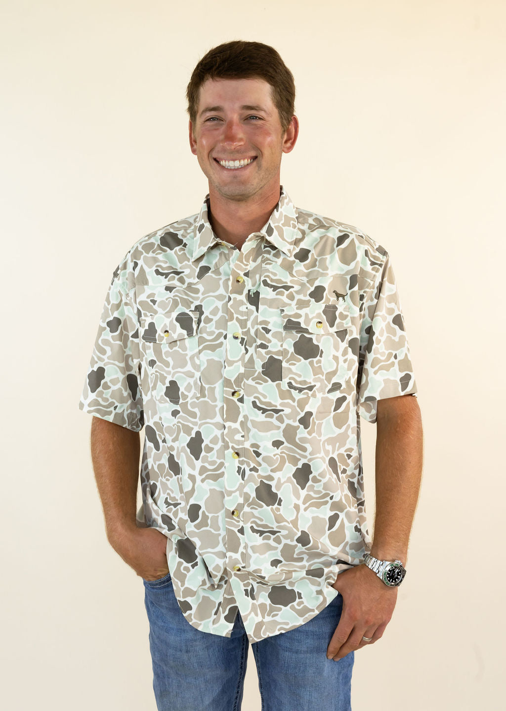 A smiling man wearing a short-sleeve button-up shirt with a beige, gray, and white camouflage pattern, paired with blue jeans, standing against a plain cream background.
