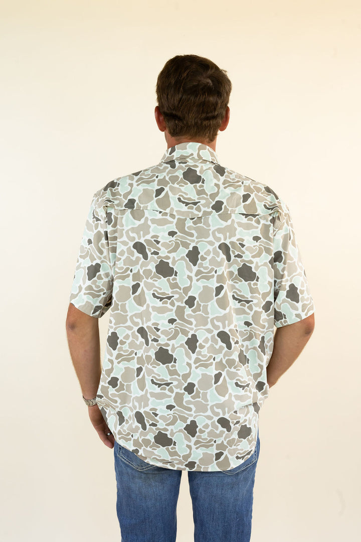 A man viewed from behind wearing a short-sleeve button-up shirt with a beige, gray, and white abstract camouflage pattern, paired with blue jeans, against a plain cream background.