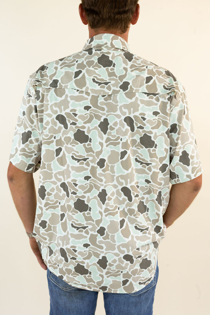 A man viewed from behind wearing a short-sleeve button-up shirt with a light beige, gray, and mint green abstract camouflage pattern, paired with blue jeans against a plain cream background.