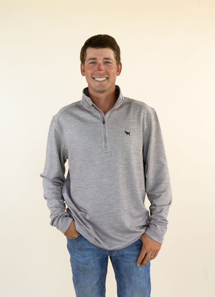 A smiling man wearing a heather brown quarter-zip pullover sweater with a small black dog logo on the left chest, paired with blue jeans, standing against a plain cream background.