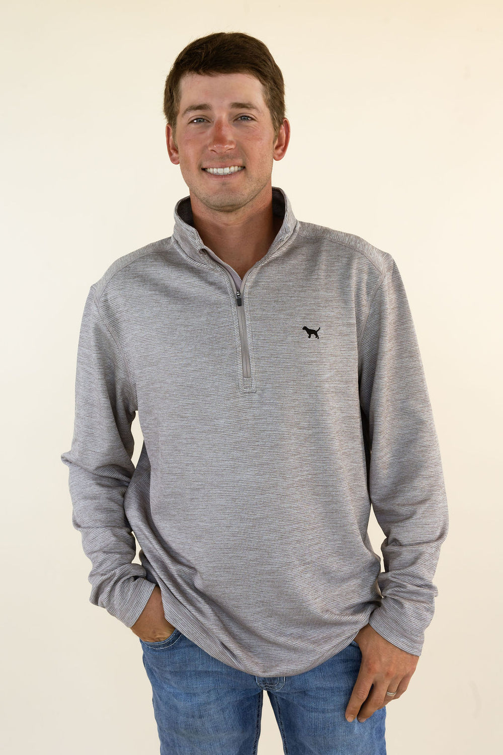 A smiling man wearing a heather brown quarter-zip pullover sweater with a small black dog logo on the left chest, paired with blue jeans, standing against a plain cream background.