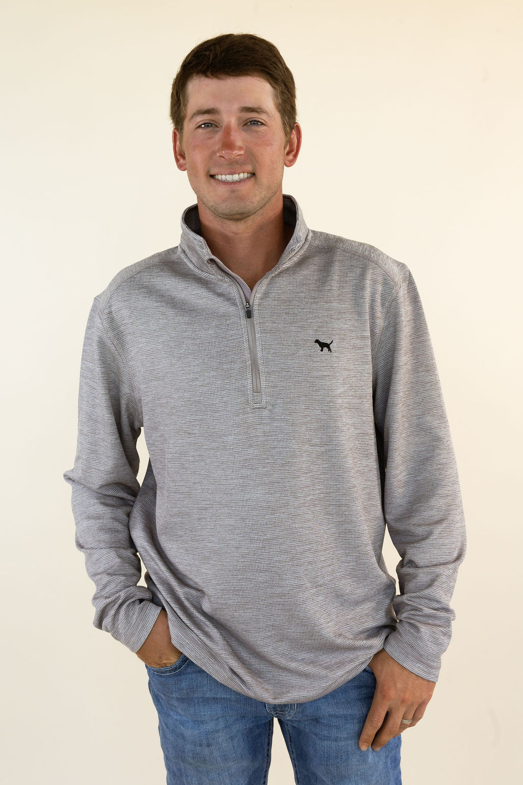 A smiling man wearing a heather brown quarter-zip pullover sweater with a small black dog logo on the left chest, paired with blue jeans, standing against a plain cream background.