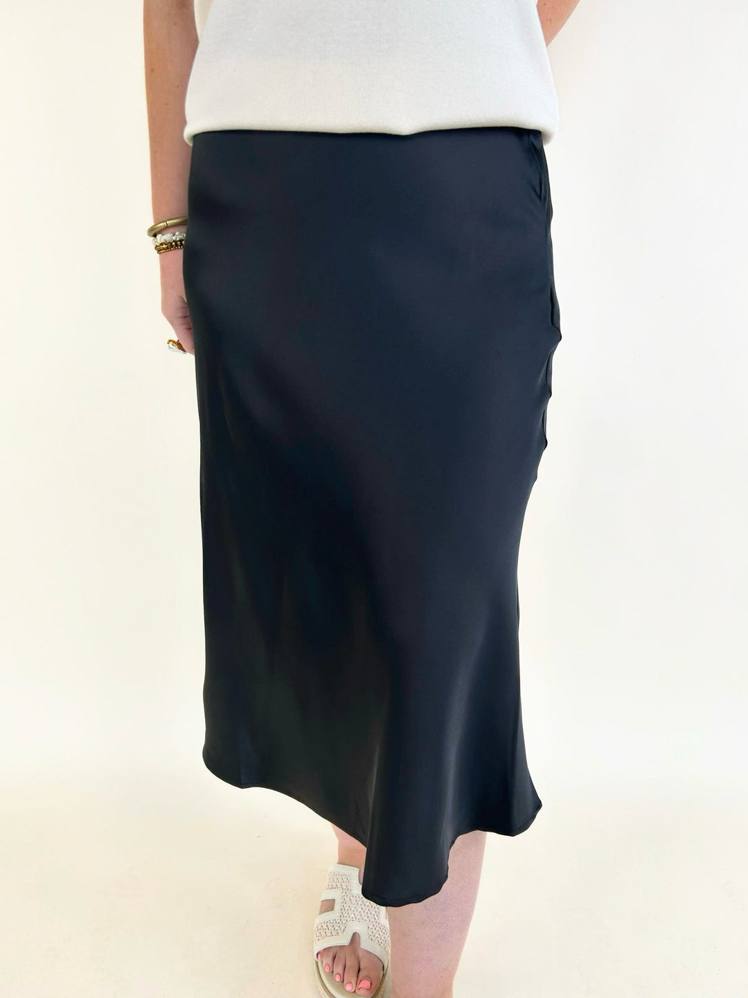 Women's black satin midi skirt with a flattering bias-cut silhouette, silky finish, and elegant everyday styling paired with a white sleeveless top.
