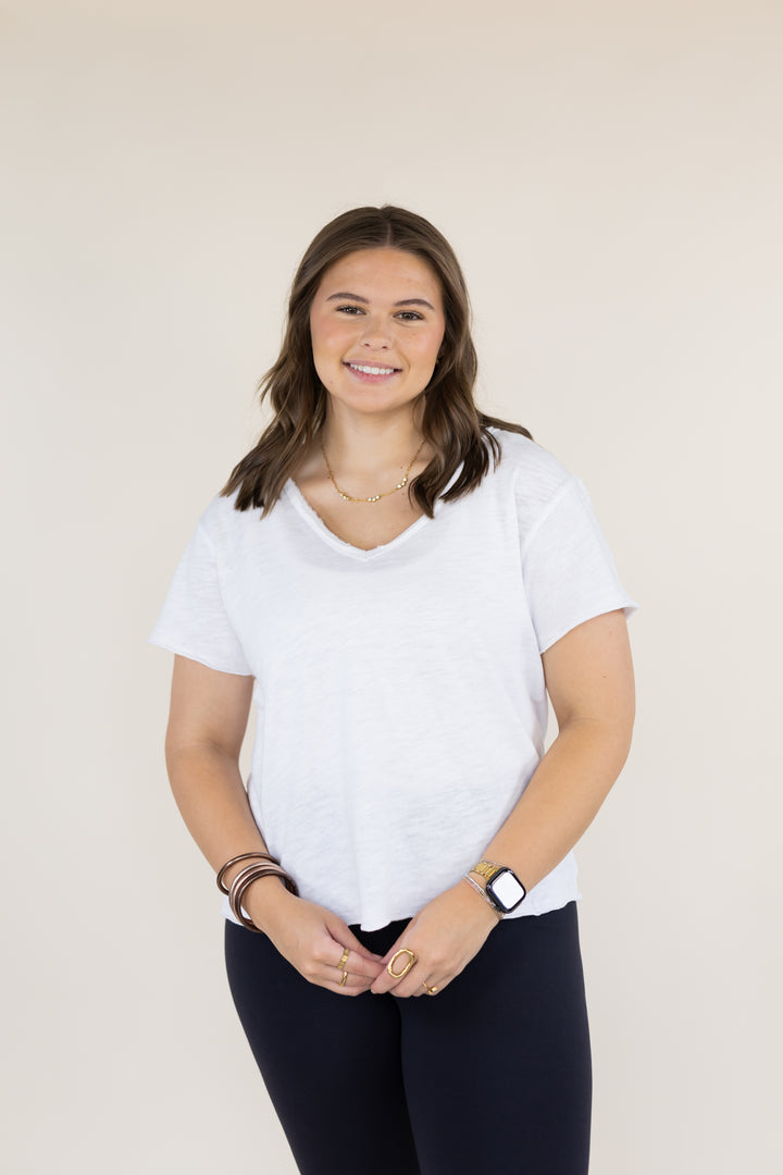 A woman with shoulder-length brown hair stands against a plain beige studio background, wearing a white short-sleeve V-neck T-shirt and dark navy pants.