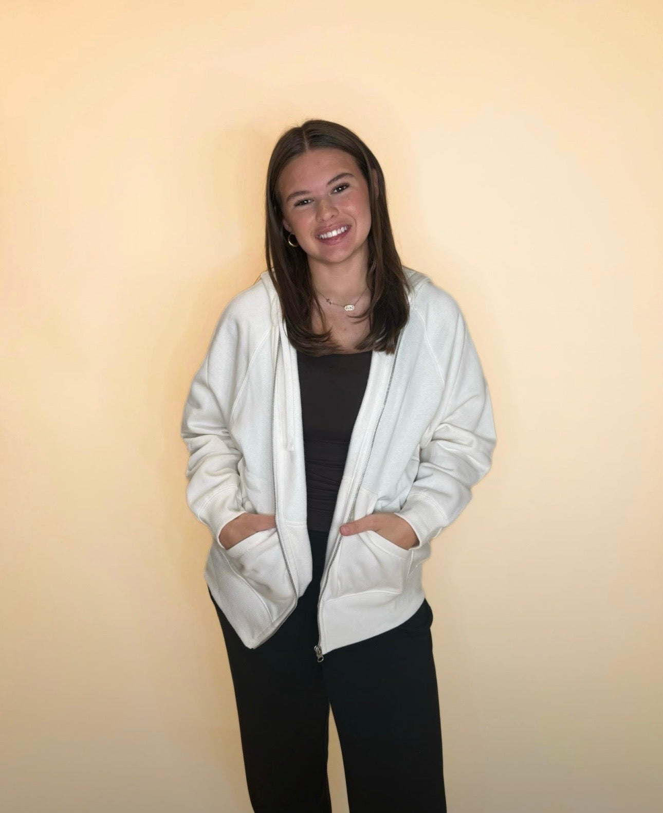 Woman wearing ivory zip-up hoodie over dark top with black pants, hands in pockets, casual cozy outfit.