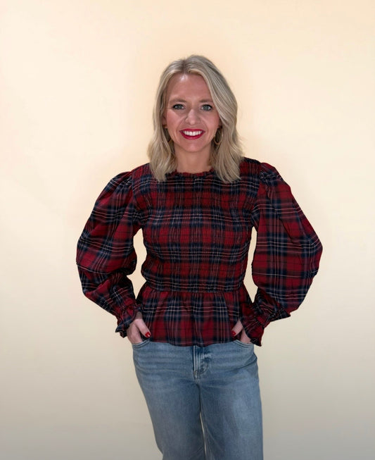 Woman wearing a red and navy plaid long sleeve top with smocked bodice and peplum hem, styled with light wash denim jeans standing against neutral background.