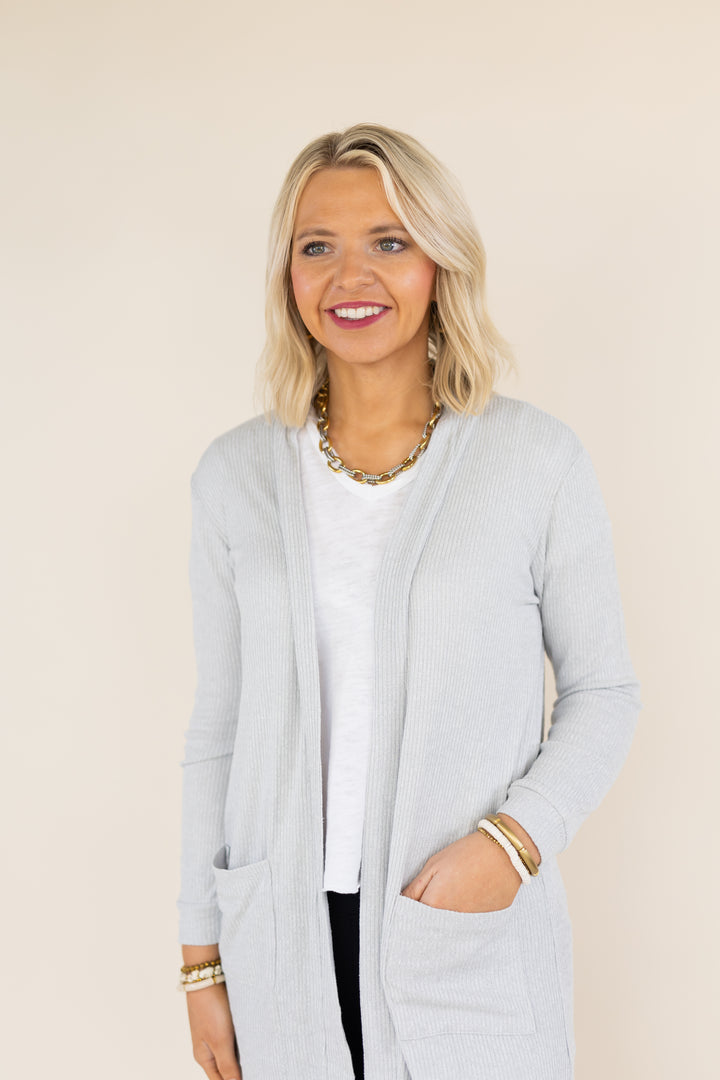 A smiling blonde woman wearing a light gray ribbed open-front cardigan over a white top, photographed against a plain beige studio background.