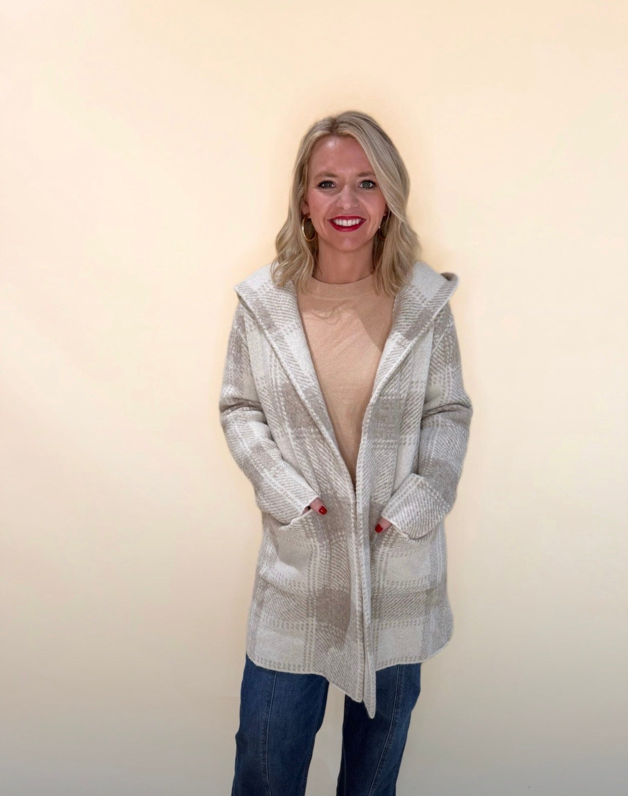 Front view of a woman wearing a taupe and cream plaid hooded cardigan layered over a beige top, paired with blue jeans, smiling with hands in pockets against a neutral background.