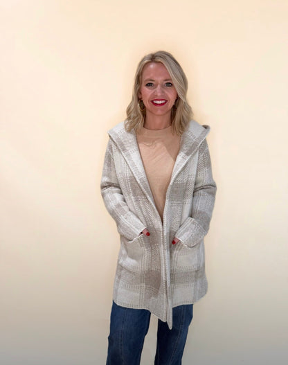 Front view of a woman wearing a taupe and cream plaid hooded cardigan layered over a beige top, paired with blue jeans, smiling with hands in pockets against a neutral background.