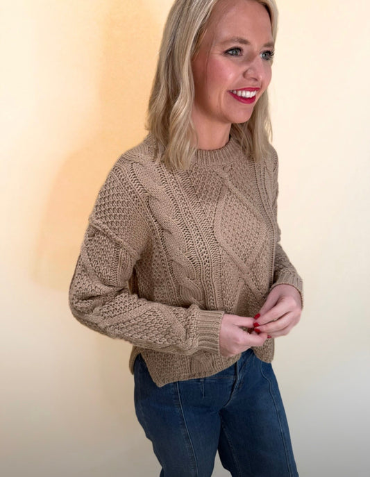 Side view of a woman wearing a tan cable knit sweater paired with high-rise blue jeans, smiling and standing against a neutral cream background.
