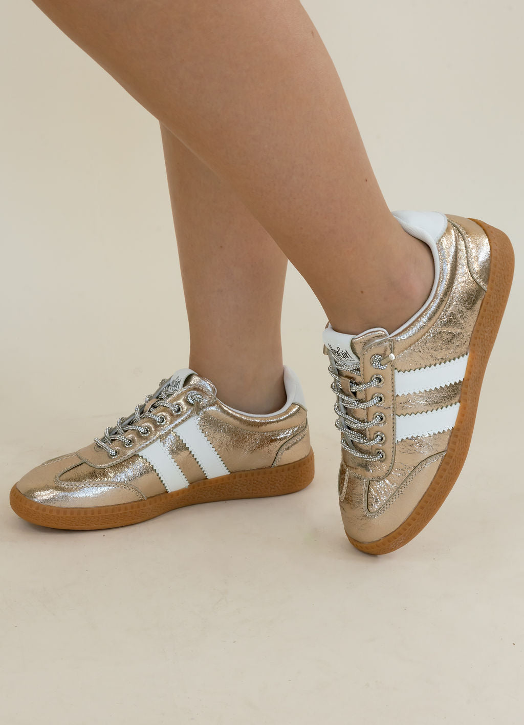 Metallic gold low-top sneakers with white side stripes and brown gum soles, shown on a model&
