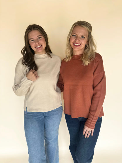 Two women wearing sweaters and jeans standing against a plain background