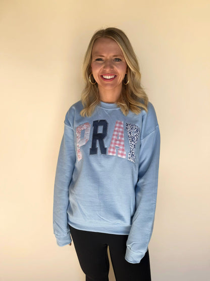 Front view of a woman in a light blue “PRAY” sweatshirt featuring mixed-pattern letters and black leggings.