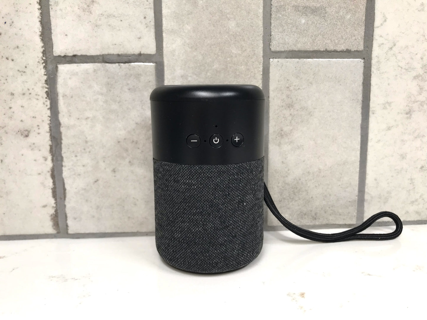 Black portable speaker with textured fabric finish on a light surface with a tiled wall background