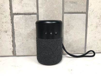 Black portable speaker with textured fabric finish on a light surface with a tiled wall background