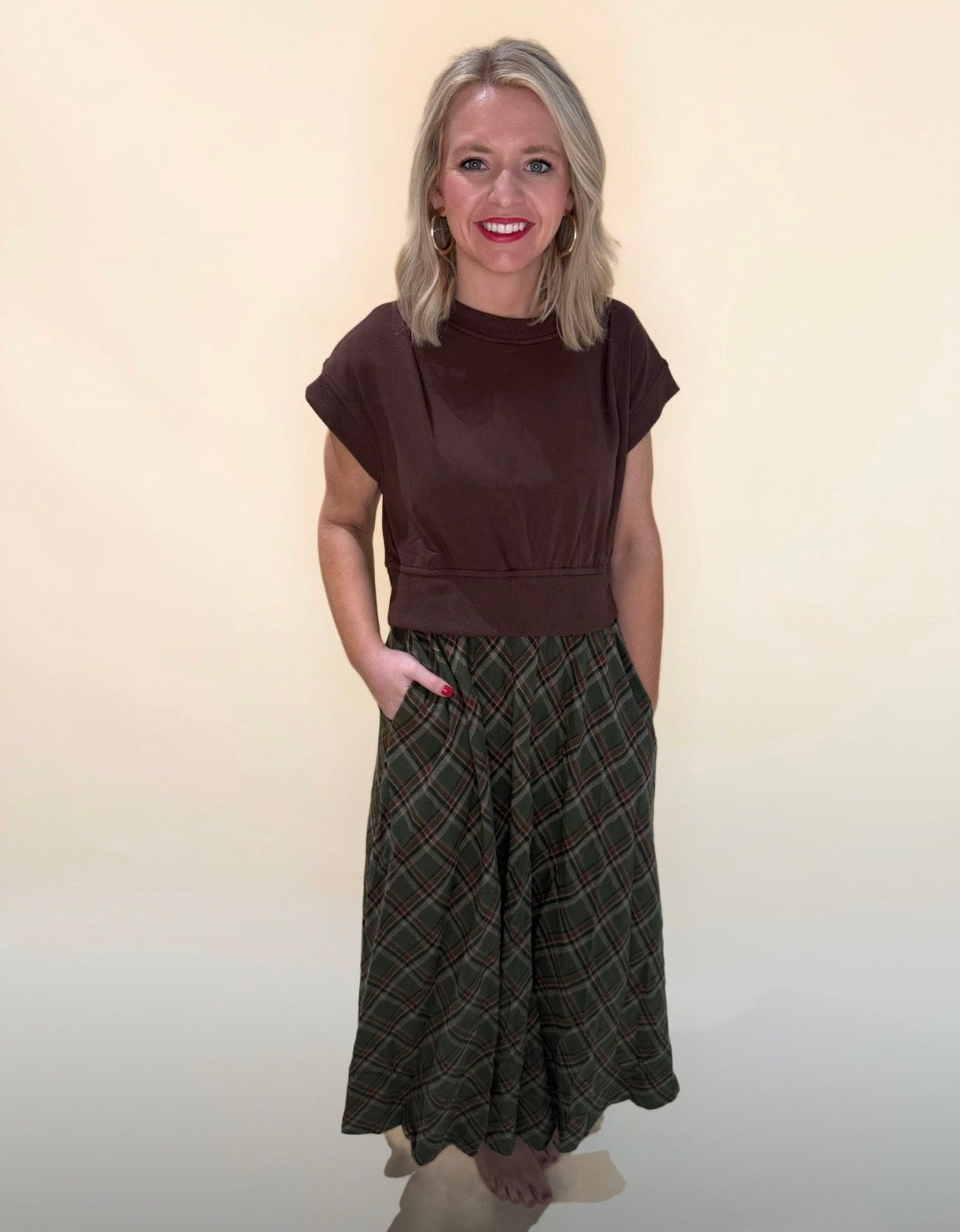 Woman wearing a chocolate brown short-sleeve top paired with a green plaid maxi skirt, hands in pockets, smiling against a neutral cream background.