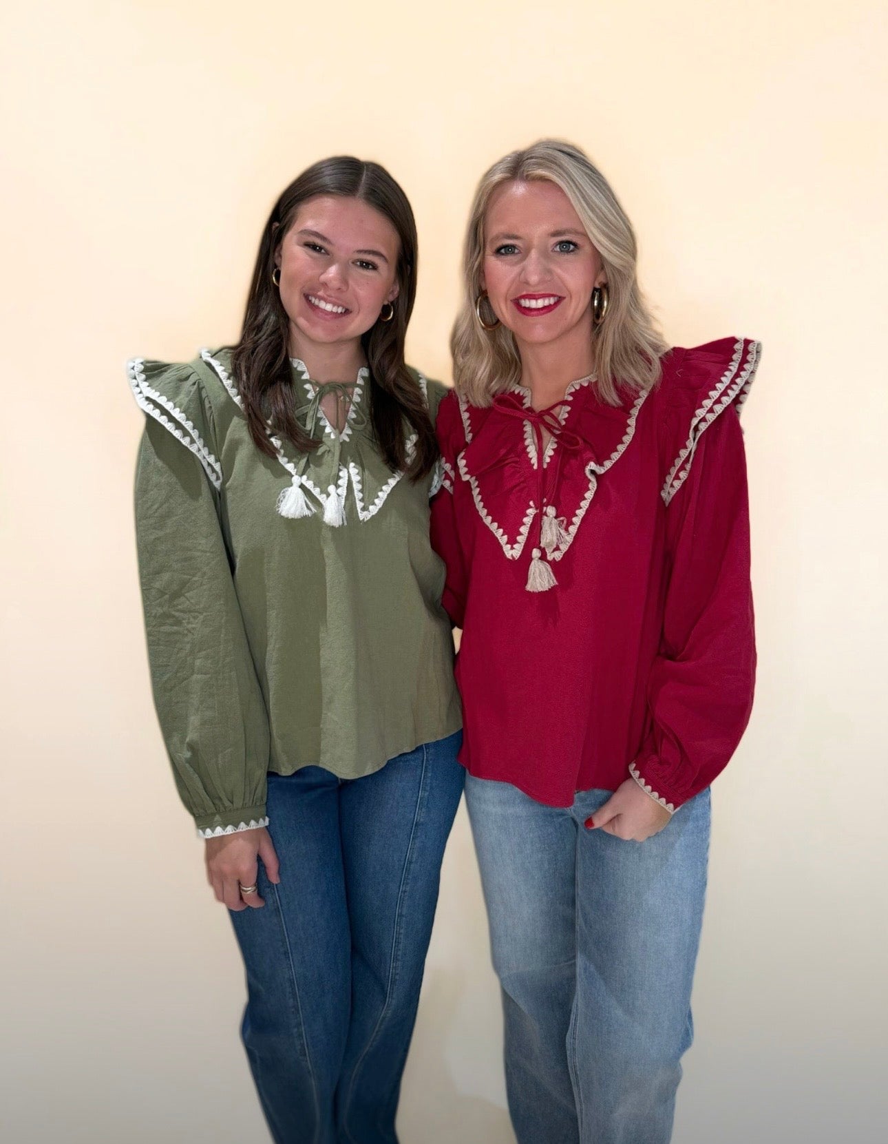 Two women wearing ruffle trim blouses with tassel ties; one in olive green and one in deep red, both styled with denim jeans against a neutral backdrop.