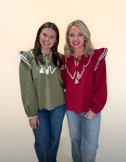 Two women wearing ruffle trim blouses with tassel ties; one in olive green and one in deep red, both styled with denim jeans against a neutral backdrop.
