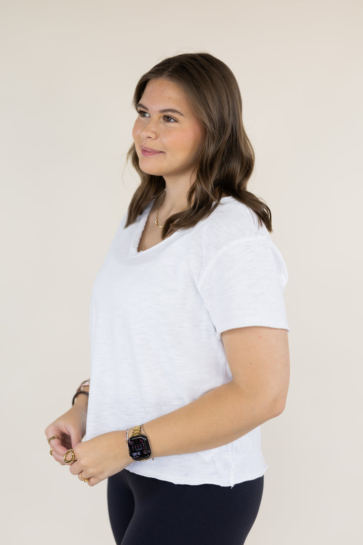 A woman wearing a white short-sleeve V-neck t-shirt and black leggings, standing against a plain beige background.