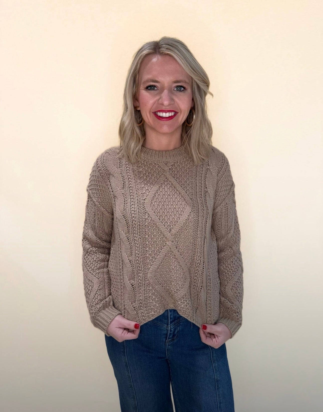Front view of tan cable knit sweater with diamond knit detailing, styled with blue jeans, model smiling with hands in pockets.