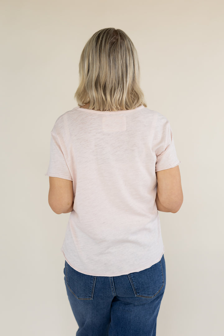A woman with blonde hair is shown from behind wearing a light pink short-sleeve t-shirt and blue jeans against a plain beige background.