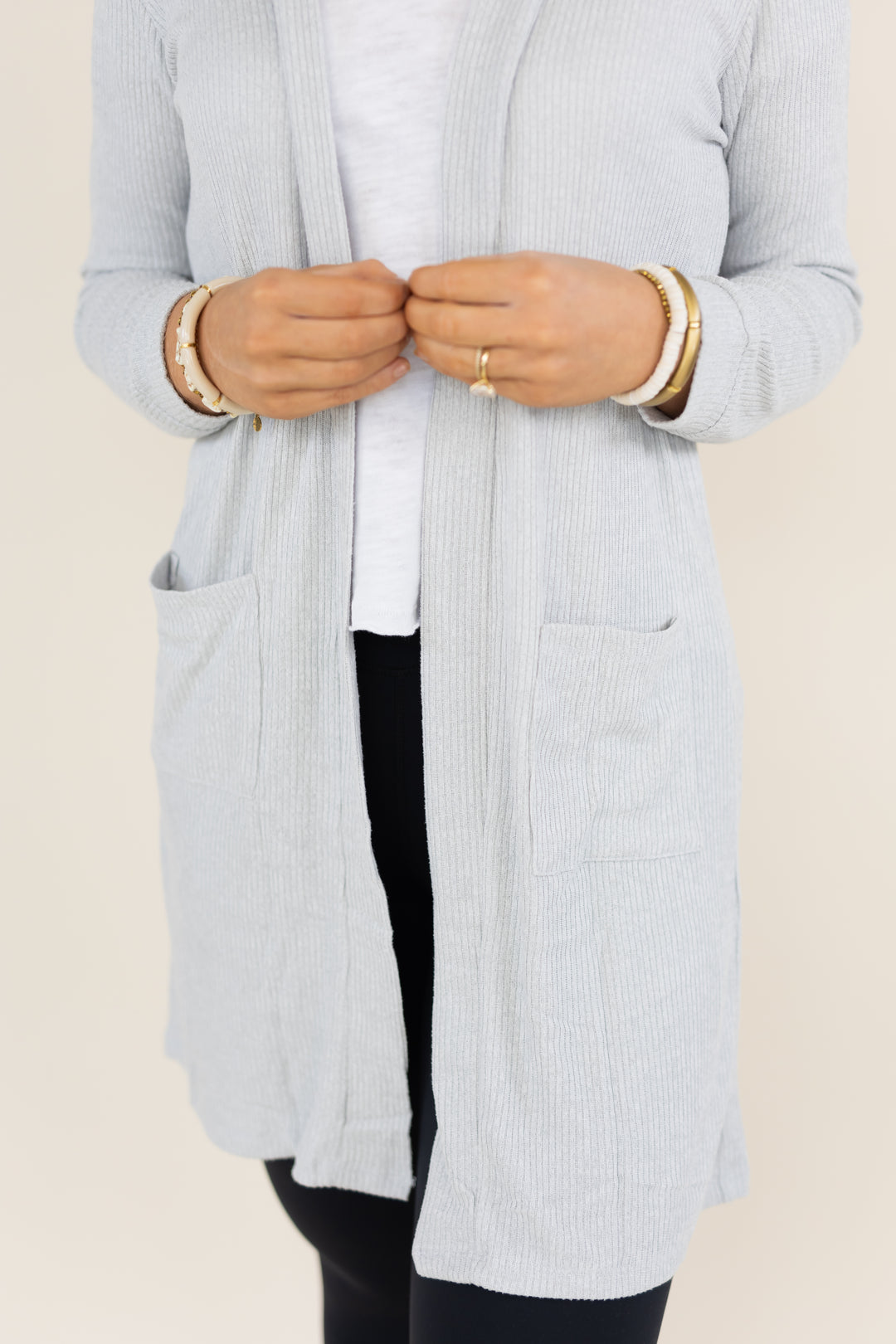 A person wearing a light gray ribbed open-front cardigan over a white top and black pants, shown from the chest down against a plain light beige background.