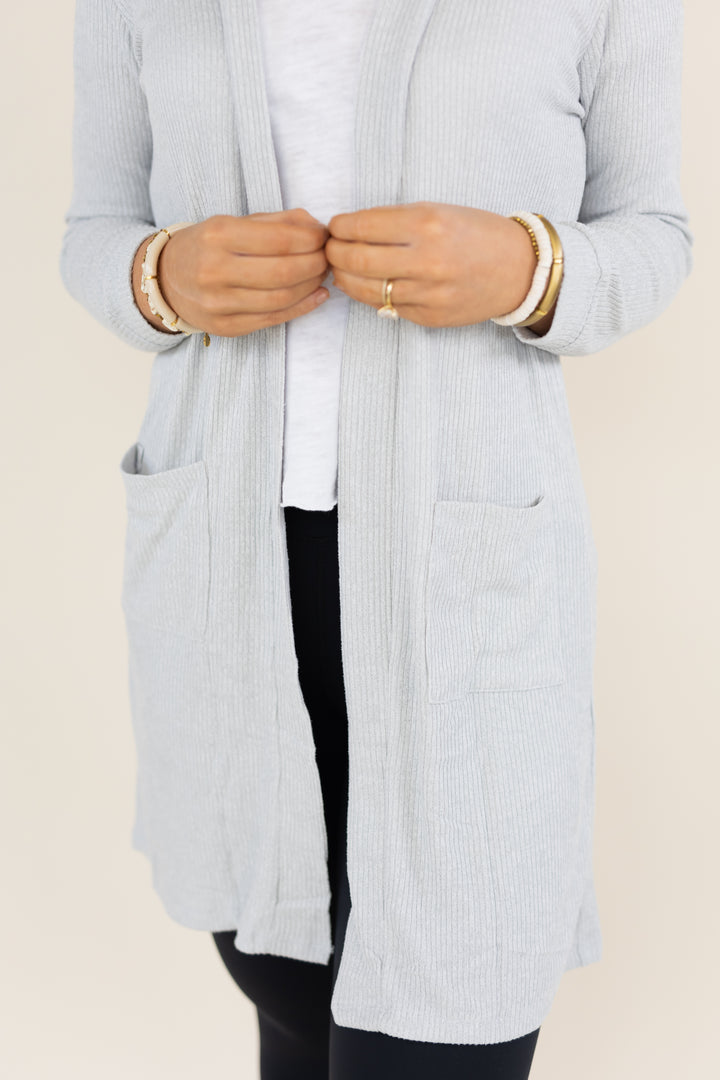 A person wearing a light gray ribbed open-front cardigan over a white top and black pants, shown from the chest down against a plain light beige background.