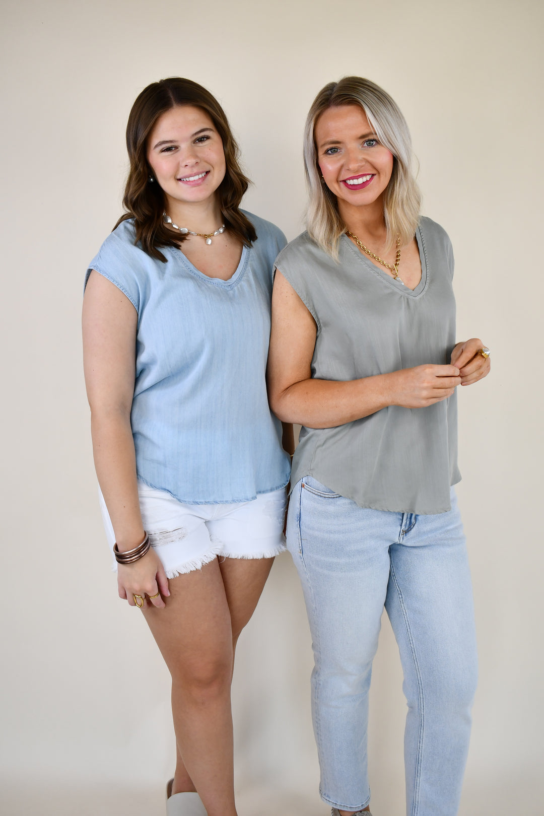 Two women wearing the Dear John Yanis V-Neck Cap Sleeve Top in Skyway Light Wash and Iceberg Green, styled with denim shorts and jeans.