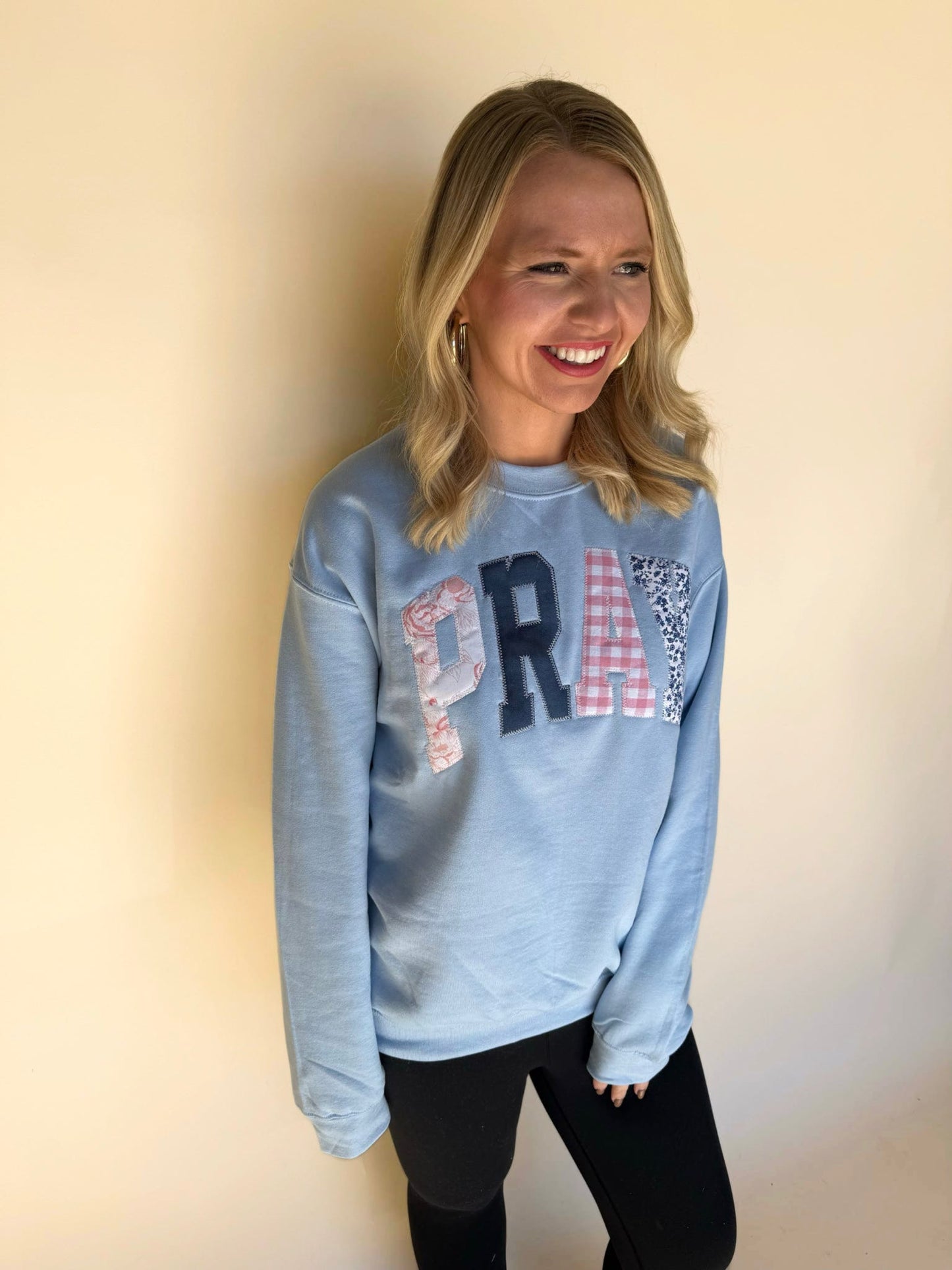 Woman smiling while wearing a light blue patchwork “PRAY” sweatshirt with black leggings, standing against a neutral background.