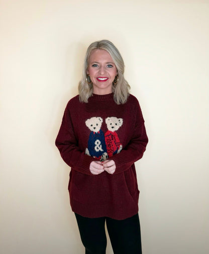 Woman wearing a burgundy oversized sweater with two teddy bear graphics and black pants, posing in front of a cream background.
