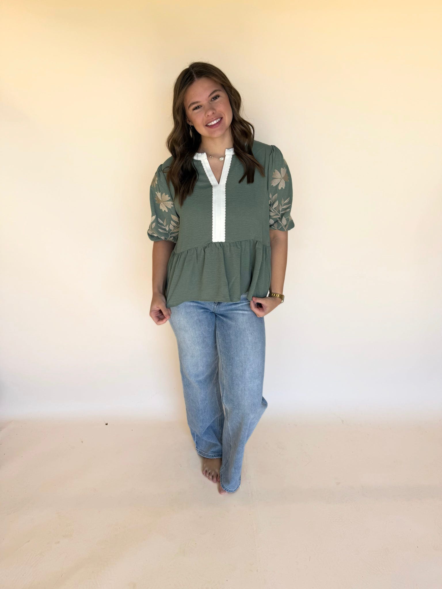 Full-body view of a woman in a sage green embroidered blouse with puff sleeves paired with light wash wide-leg jeans.