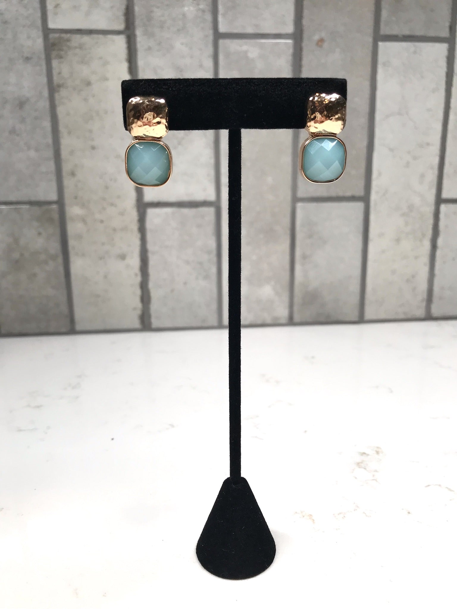 Gold hammered stud earrings with faceted light blue drop stones on a black display stand.