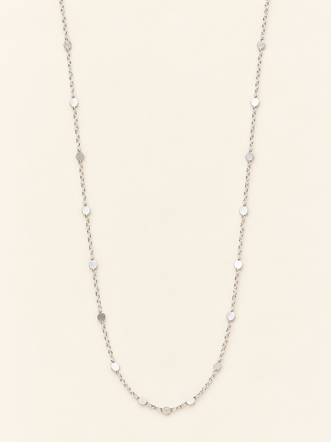 Long silver flat disc station chain necklace arranged on a cream studio background, featuring delicate chain links and evenly spaced polished round accents.