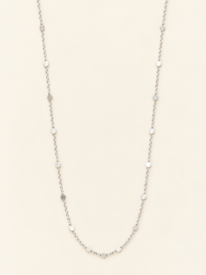Long silver flat disc station chain necklace arranged on a cream studio background, featuring delicate chain links and evenly spaced polished round accents.