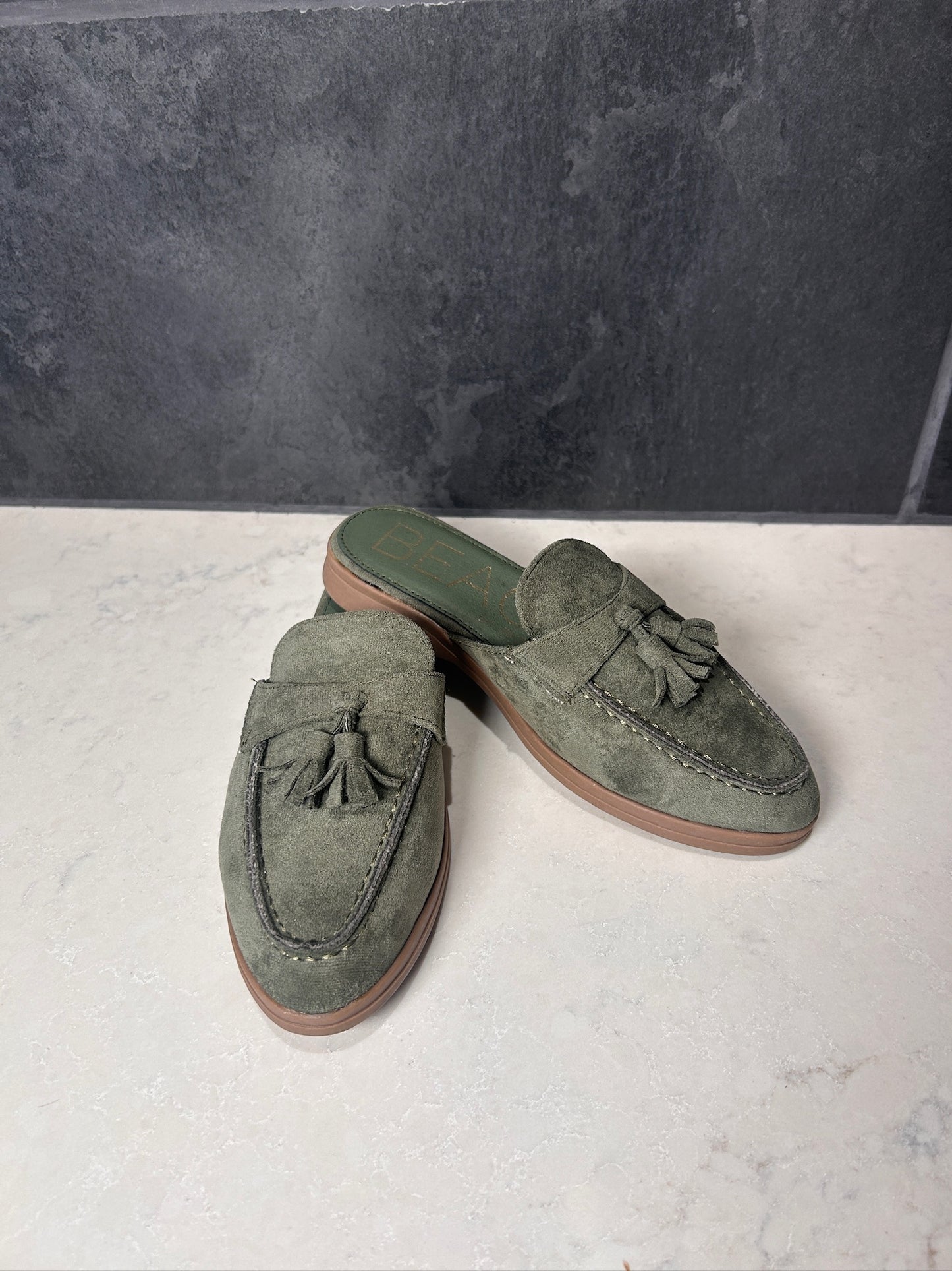 Pair of green loafers with tassels on a light gray surface with a dark gray background