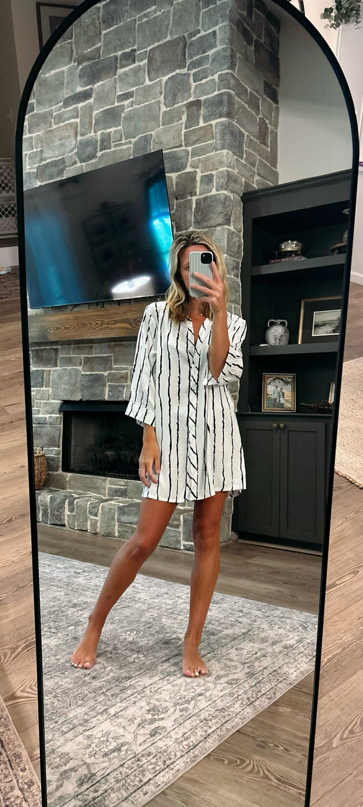A woman takes a mirror selfie in a living room wearing a white short-sleeve button-up shirt dress with thin black vertical stripes. The background features a gray stone fireplace, a black entertainment cabinet, and a light-colored rug.