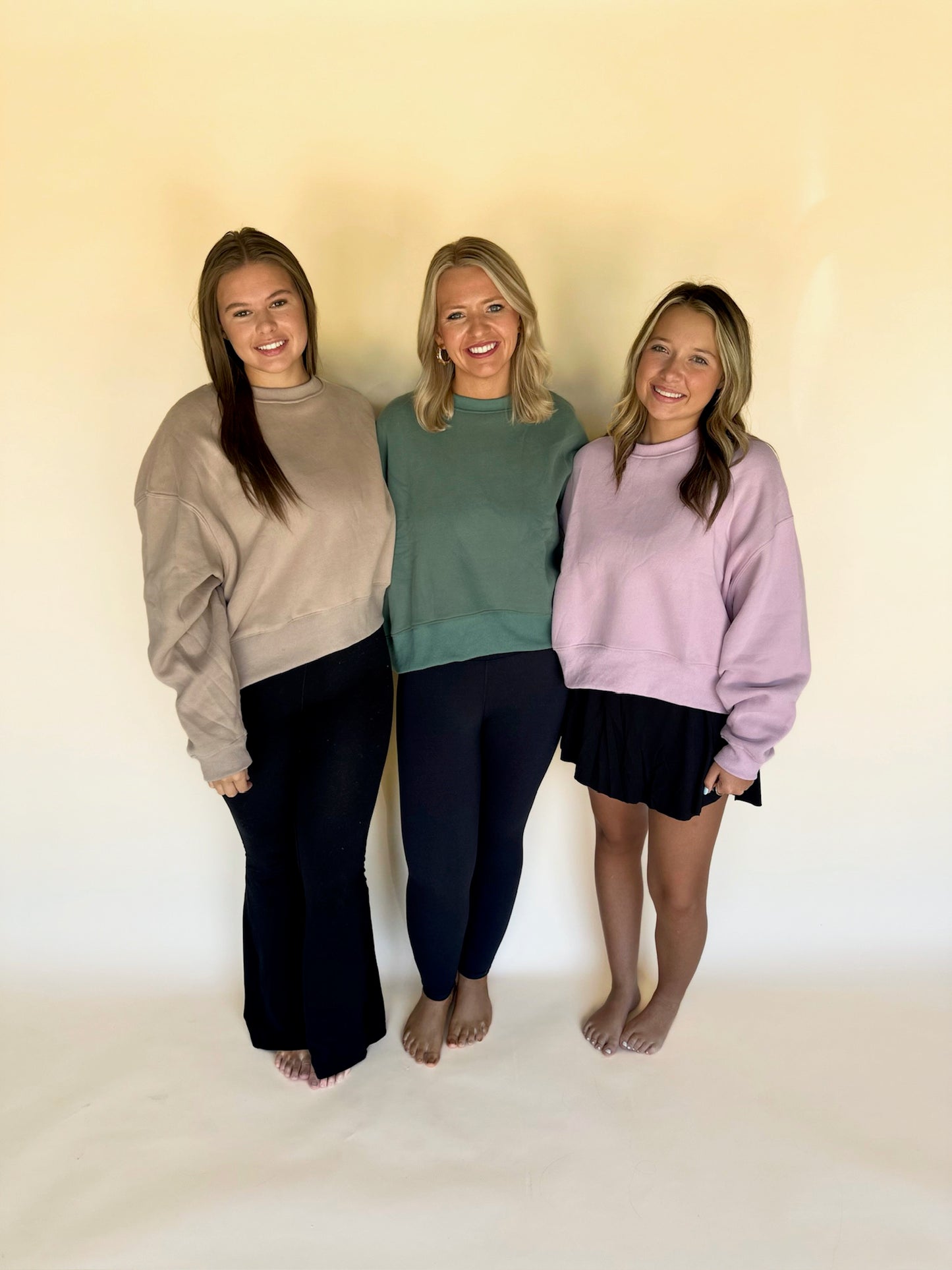 Three women standing together wearing casual outfits against a plain background