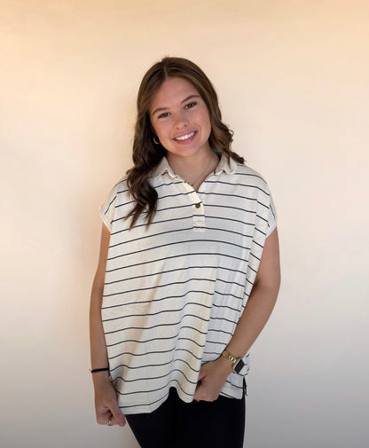“Young woman wearing a loose-fit cream and black striped collared top, smiling and standing against a neutral backdrop. 