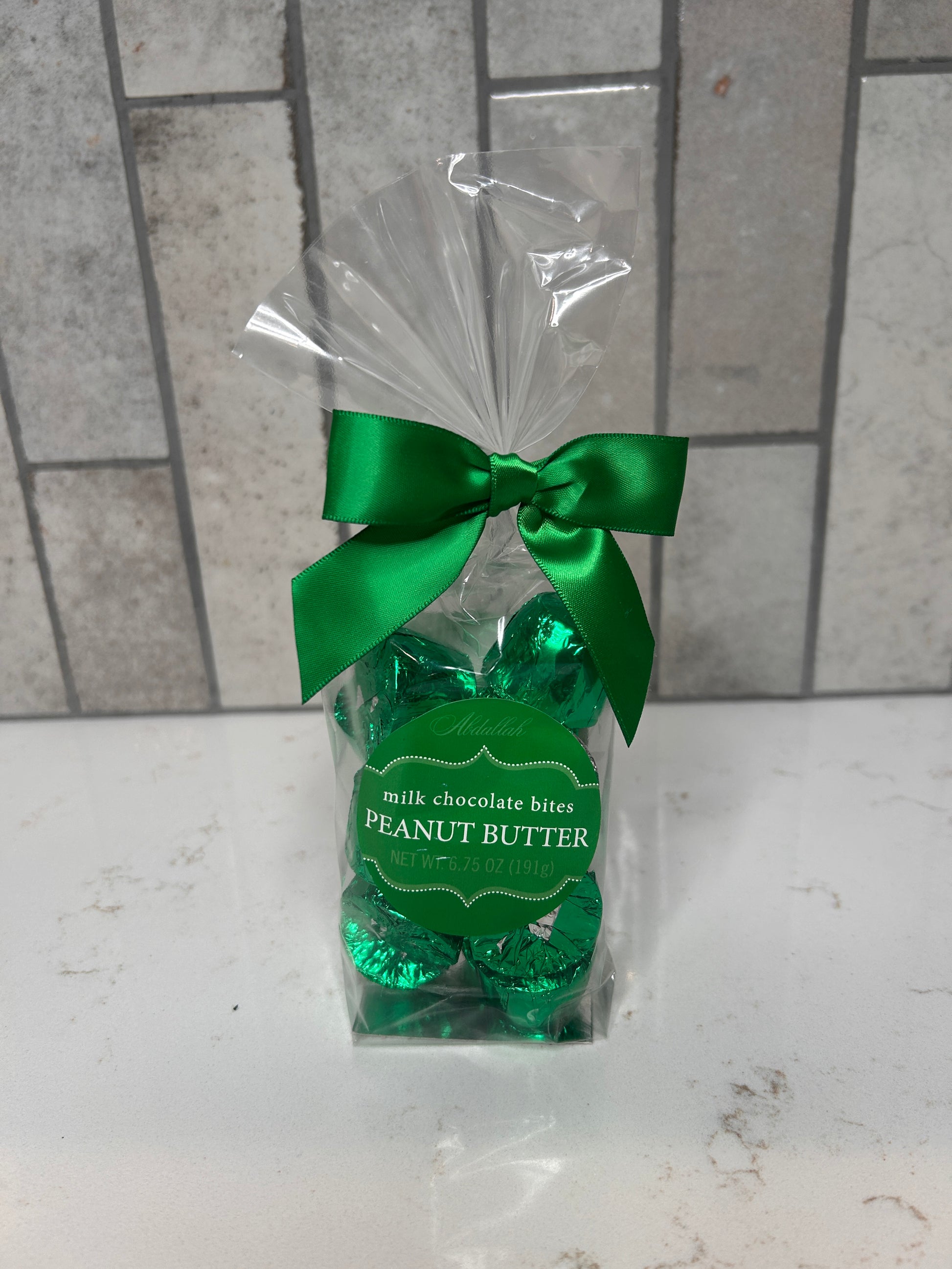 A clear gift bag filled with green-foil wrapped milk chocolate peanut butter bites, tied with a green satin bow, displayed on a white countertop with a gray tile background.